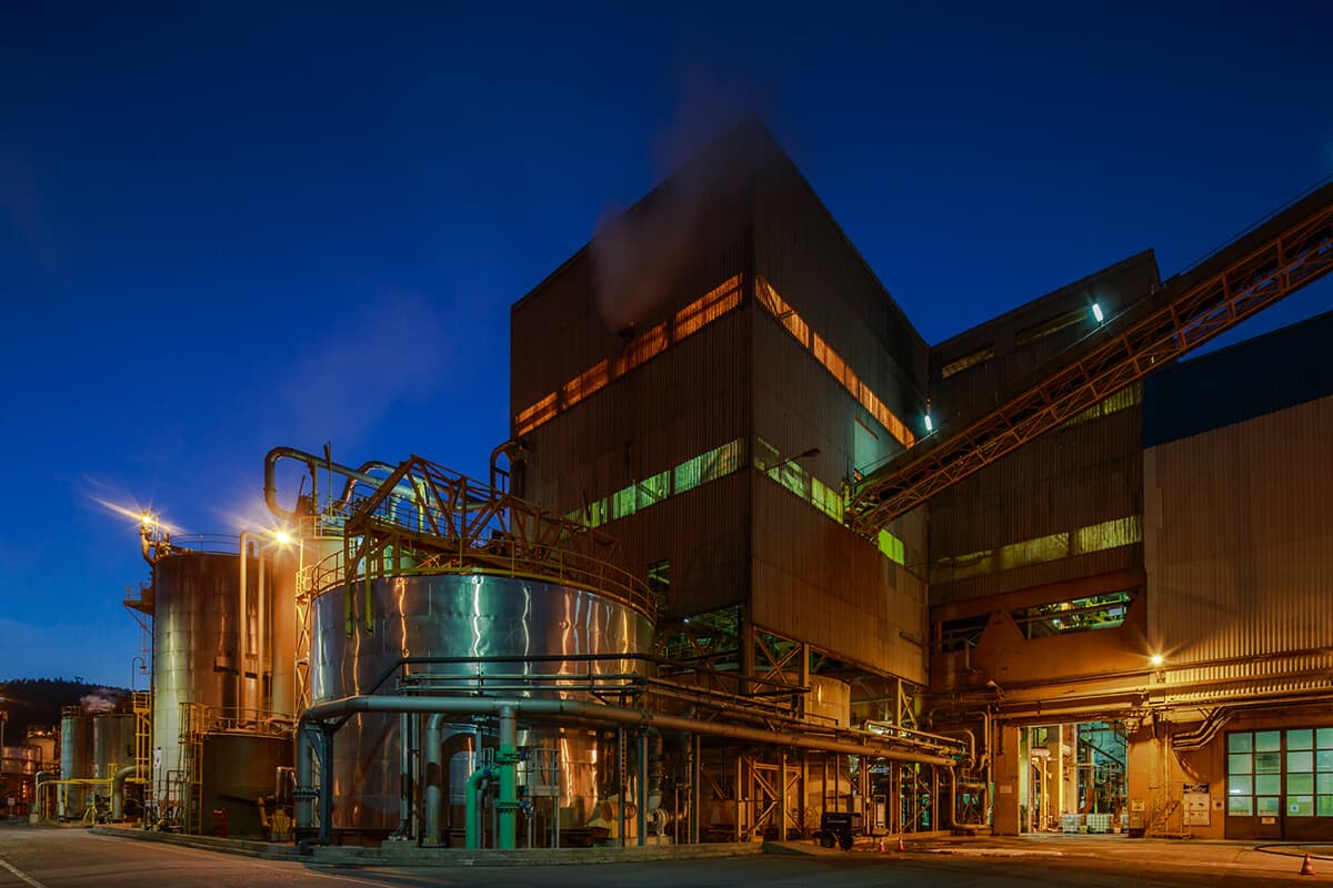 Planta de celulosa iluminada de noche, mostrando grandes estanques de almacenamiento metálicos, tuberías de proceso y estructuras industriales bajo un cielo crepuscular.