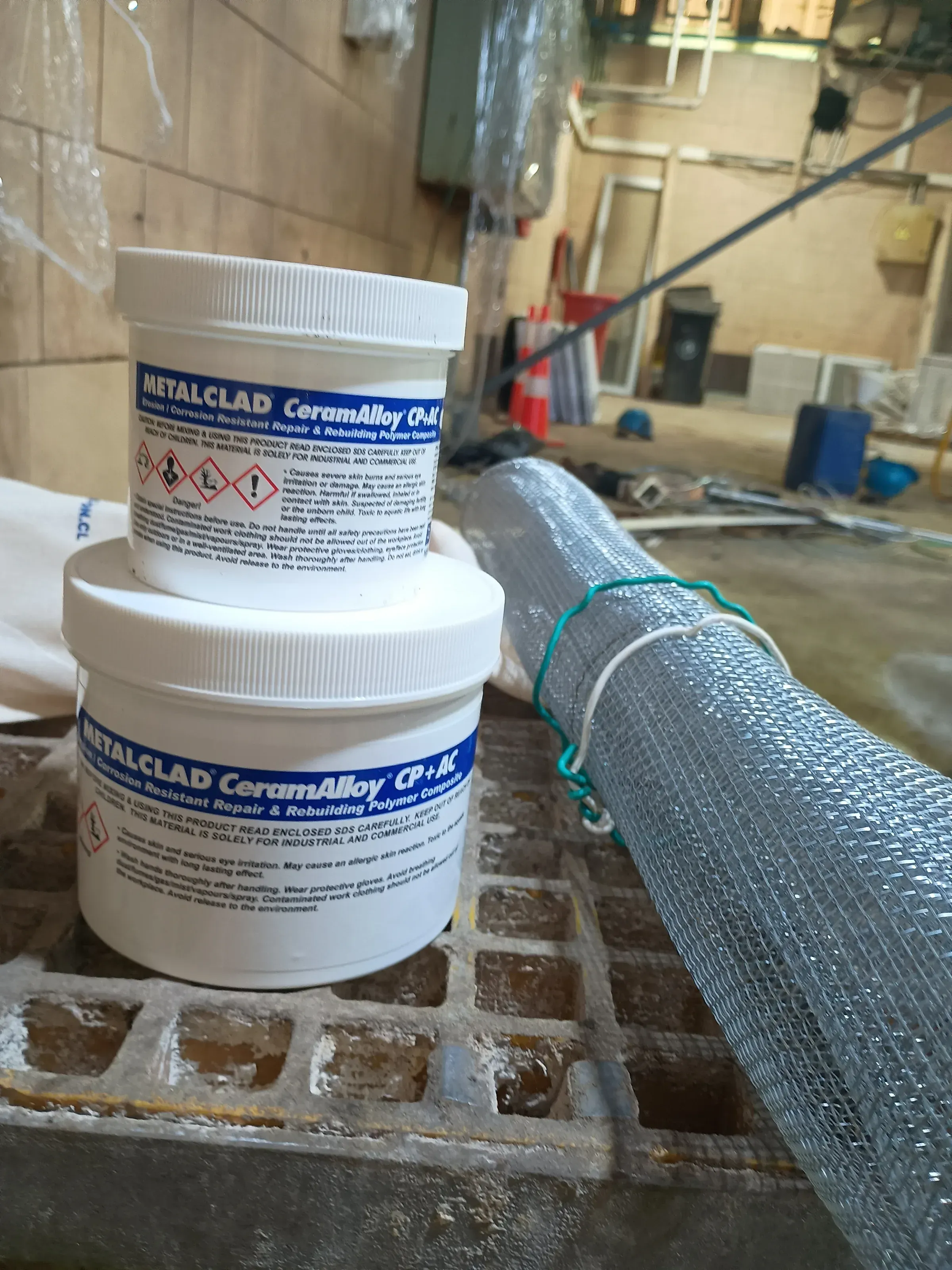 Dos envases de compuesto polimérico METALCLAD CeramAlloy CP+AC y malla metálica de refuerzo sobre rejilla industrial en área de mantenimiento.