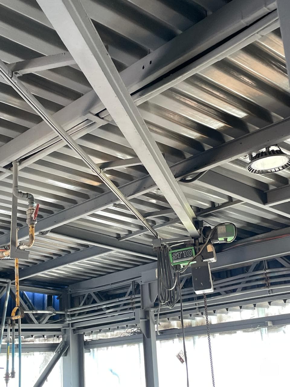 Estructura metálica de techo industrial con vigas de acero y cubierta acanalada protegidas con recubrimiento gris anticorrosivo, junto a polipasto eléctrico y tuberías.