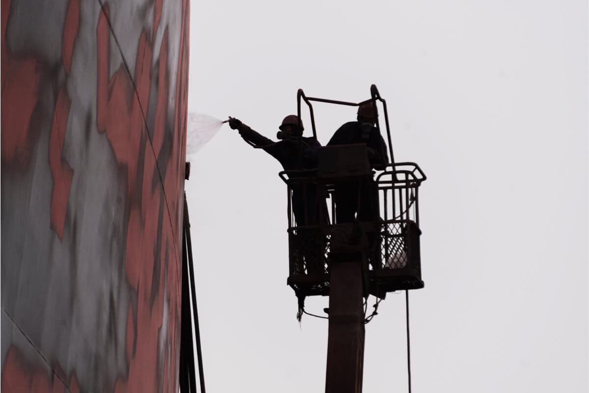 Operarios en plataforma elevadora aplicando recubrimiento protector por pulverización sobre el casco de un buque durante trabajos de mantenimiento marino.