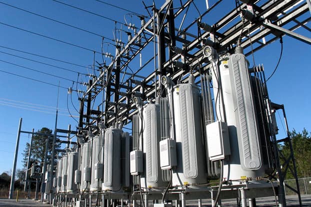 Fila de transformadores eléctricos en una subestación de energía exterior, expuestos a la intemperie y corrosión ambiental bajo un cielo despejado.