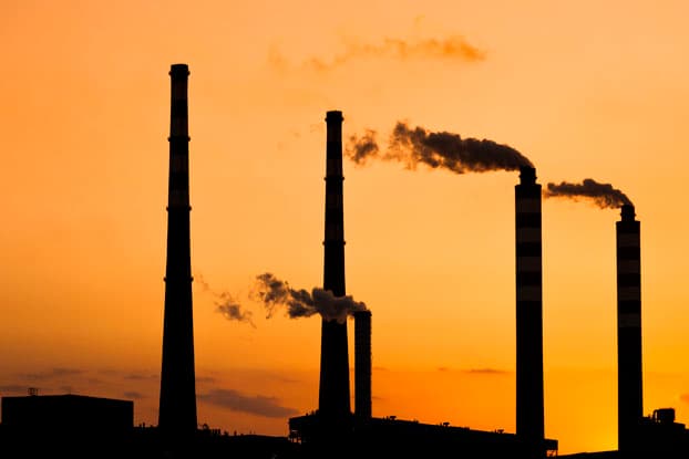 Siluetas de chimeneas industriales emitiendo emisiones contra un cielo de atardecer anaranjado en una planta de generación de energía.