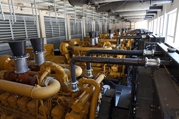 Fila de motores generadores industriales amarillos en una sala de máquinas, mostrando sistemas de tuberías, escapes y conductos de ventilación en una planta de energía.