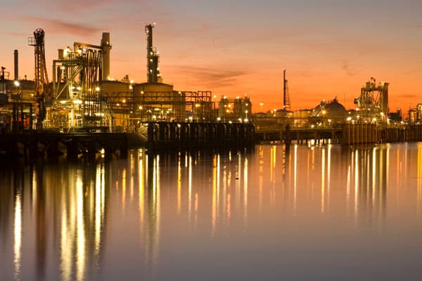 Planta de tratamiento de aguas residuales e instalaciones industriales junto al agua durante el atardecer, mostrando tanques, tuberías y luces reflejadas.