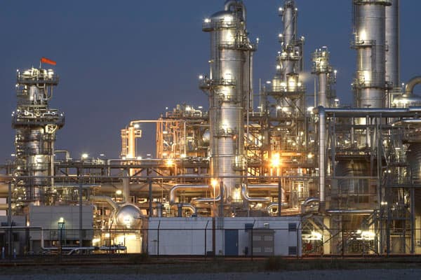 Complejo industrial o refinería iluminada al atardecer, mostrando columnas de destilación, tuberías de acero y estructuras metálicas complejas para el procesamiento de fluidos.