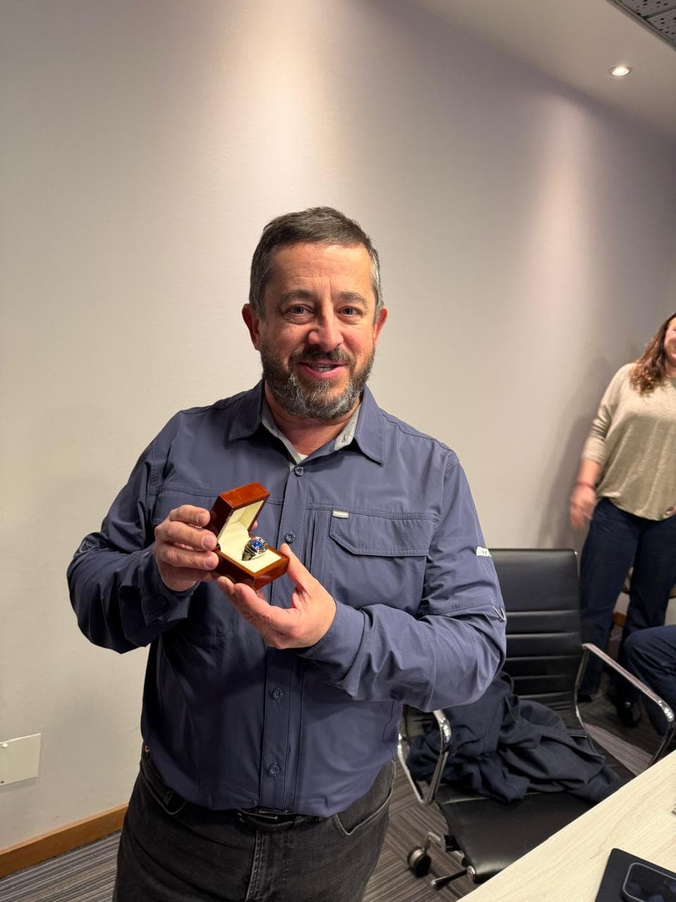 Hombre sonriente con camisa azul sosteniendo una pequeña caja de madera abierta que contiene un anillo de reconocimiento en una oficina.