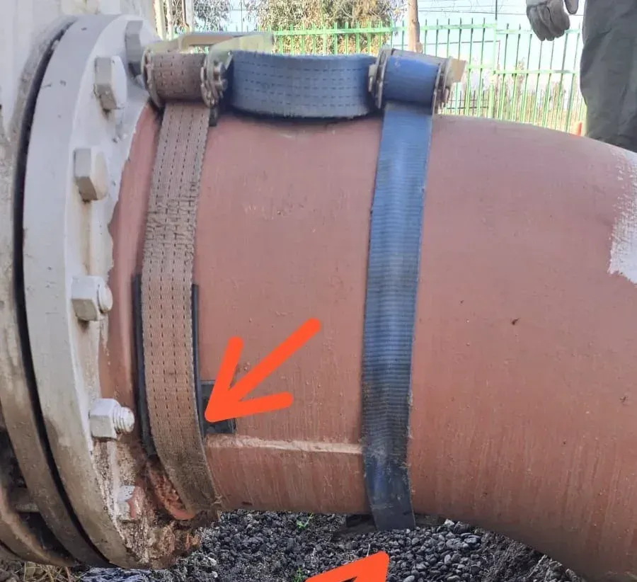 Tubería de gran diámetro con brida metálica y correas de sujeción temporales, mostrando puntos de filtración señalados con flechas rojas sobre terreno húmedo.